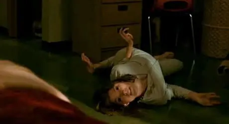 A woman lies on the floor in a distressed pose, looking directly at the viewer, with one arm positioned awkwardly behind her and an expression of fear or anguish.